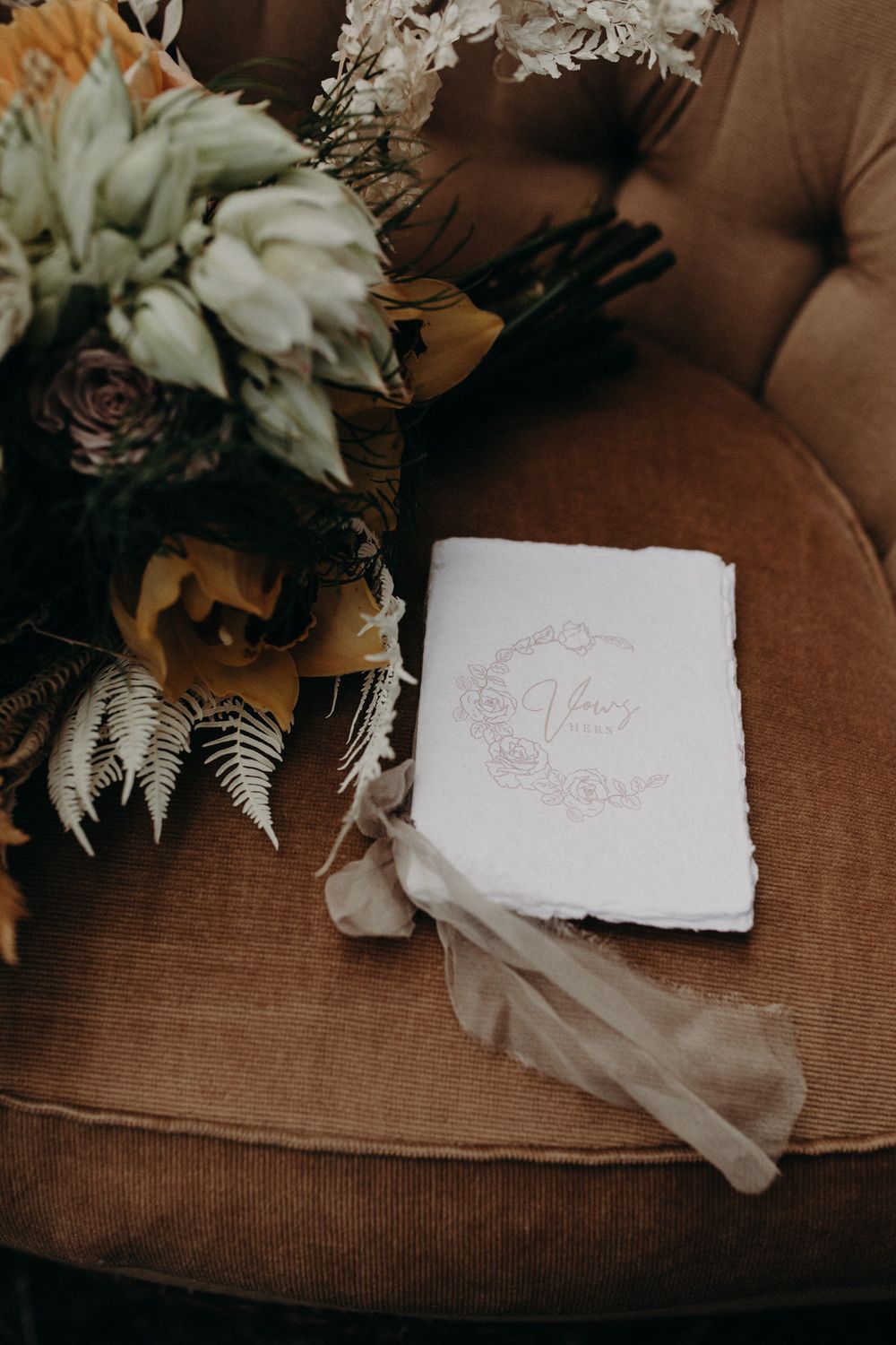 Wedding stationery and floral arrangement on brown velvet sofa.