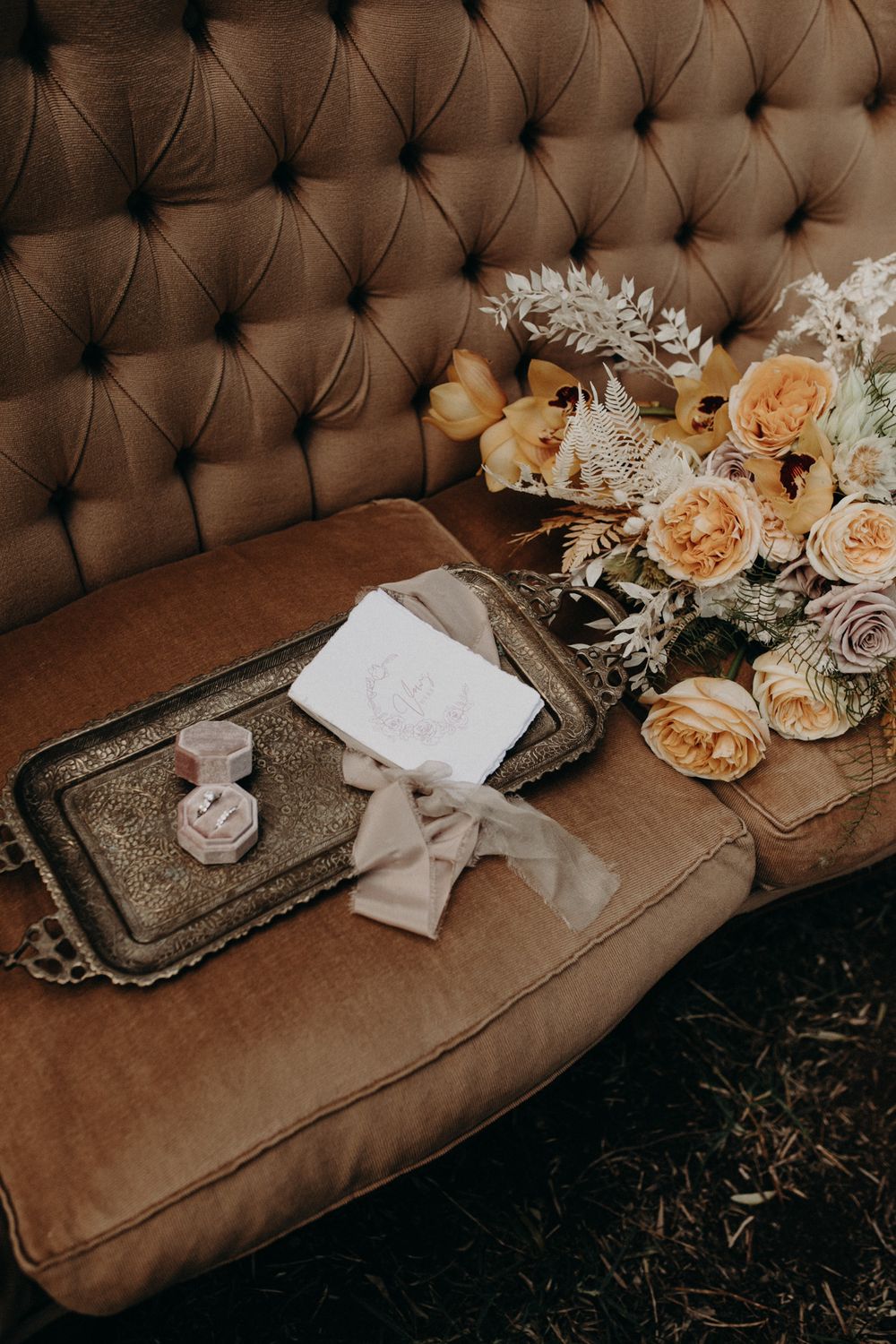 Wedding details with floral arrangement and stationery on vintage brown tufted couch.