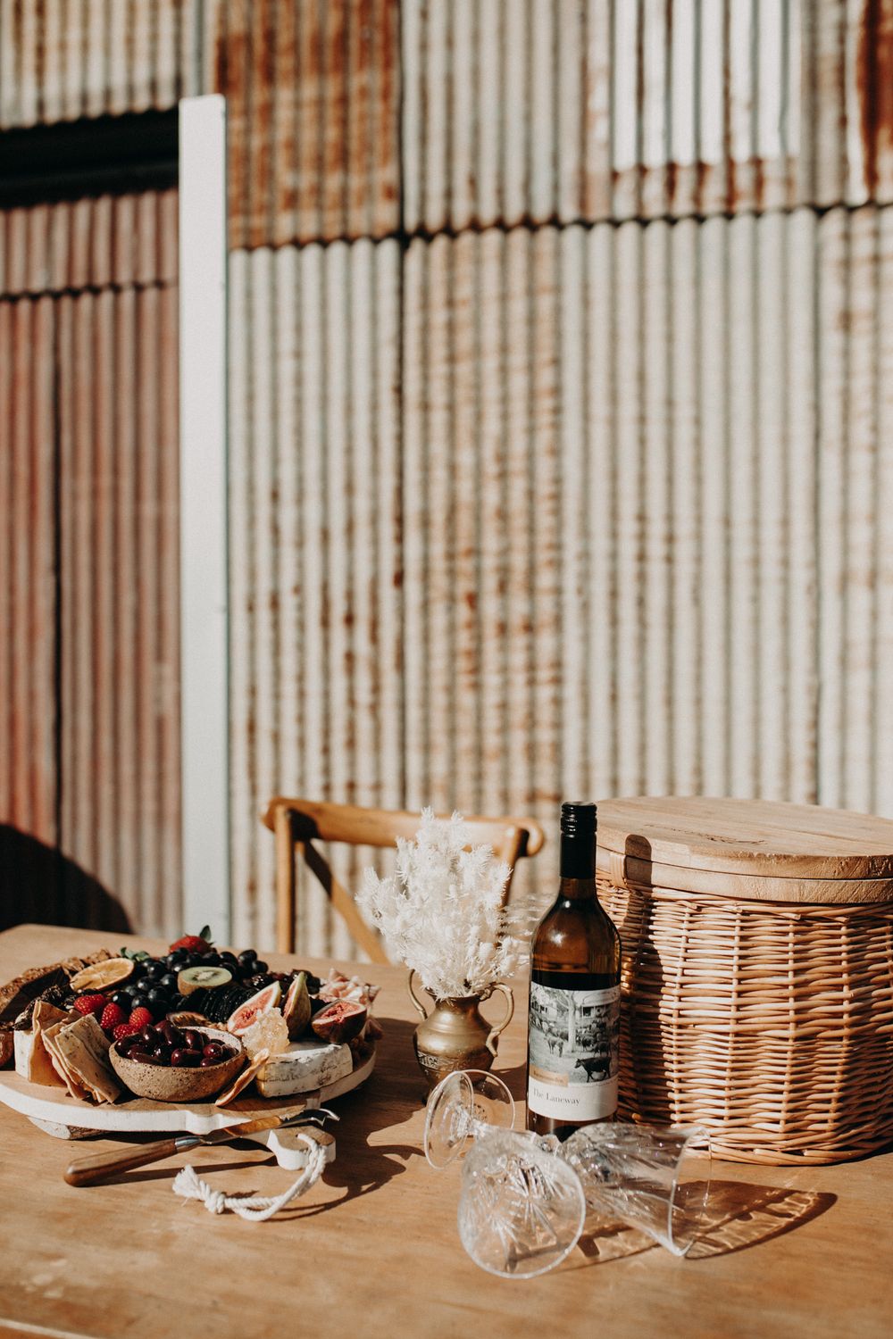 Vintage rustic table setting with wine bottle and wicker basket against corrugated metal wall.