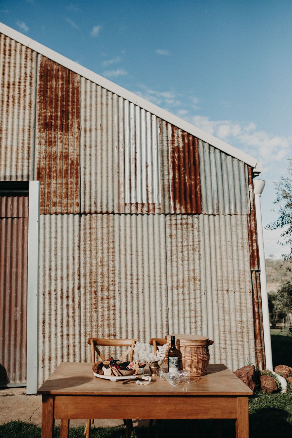 Rustic wooden table set against weathered corrugated metal barn wall under blue sky.