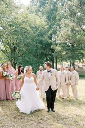 Wedding party in blush pink dresses and cream suits poses together on a sunny outdoor lawn.