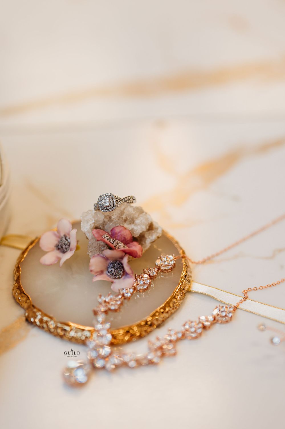 Elegant bridal jewelry flat lay with diamond ring, rose gold necklace, and pink floral accents.