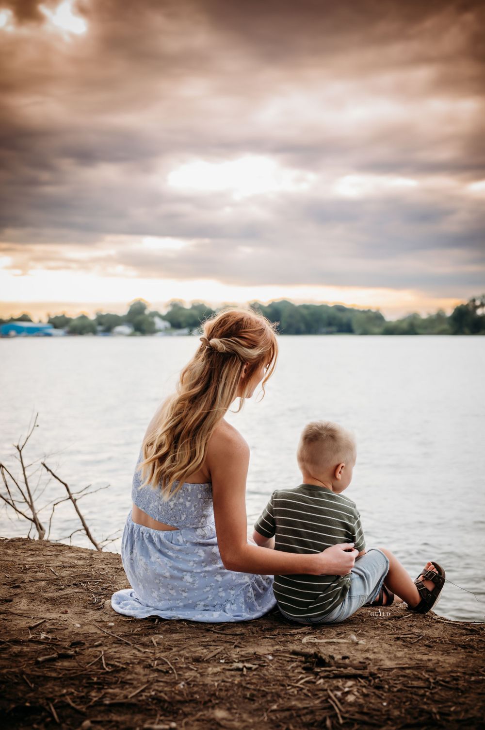 Mother and young son sitting together on a lakeside cliff watching a dramatic sunset sky.