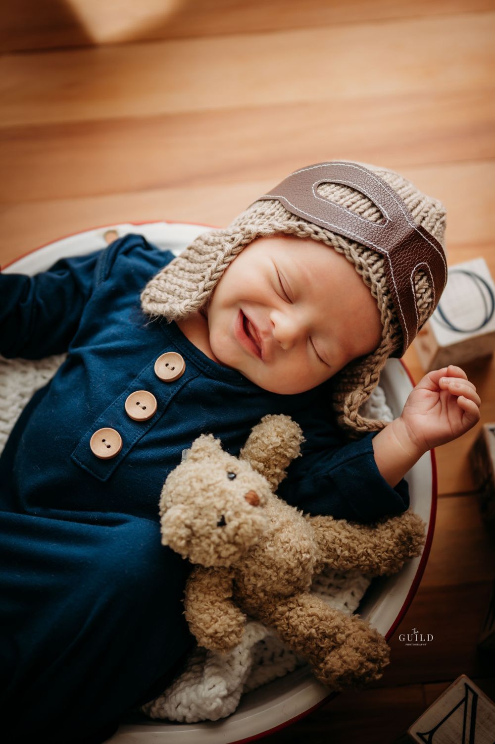 Smiling newborn baby in aviator hat and blue romper sleeping with a teddy bear in a bowl.