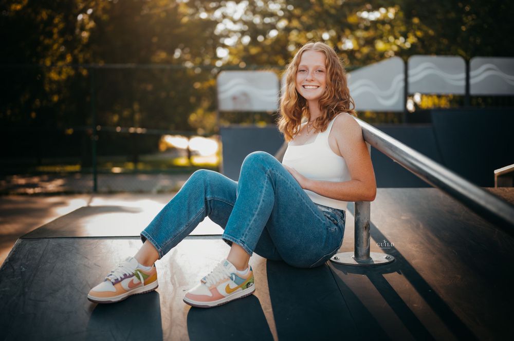 Smiling teenage girl in white tank top and jeans sitting on a skate ramp in golden sunlight.