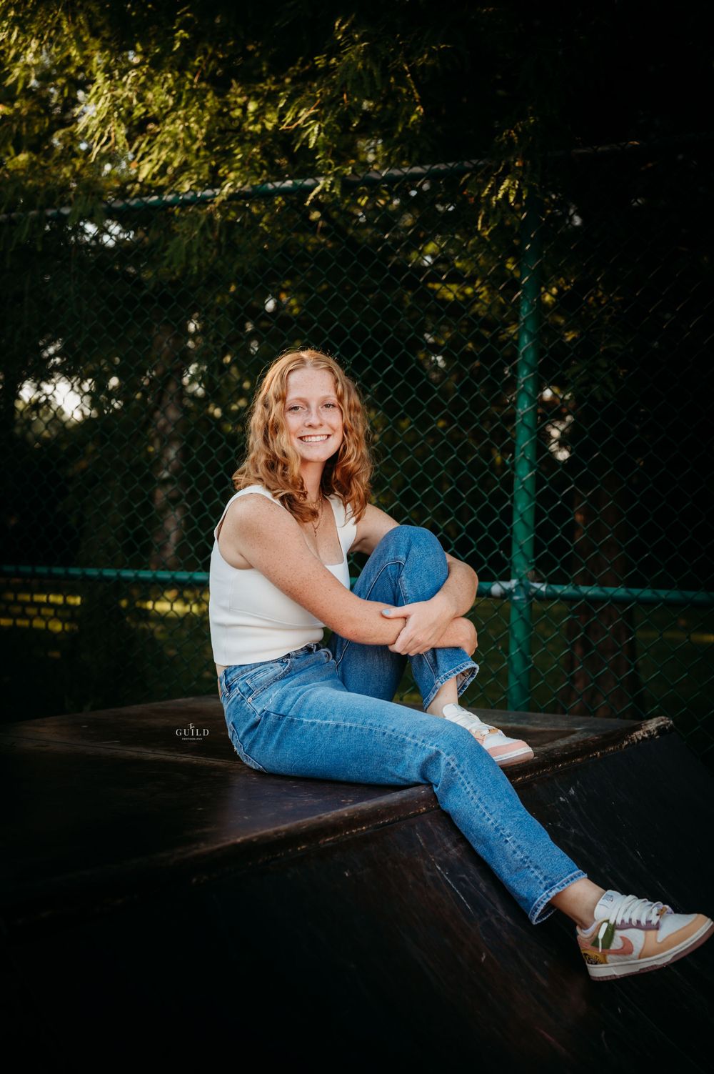 Smiling teenage girl in white tank top and jeans sitting on a trampoline surrounded by green trees.