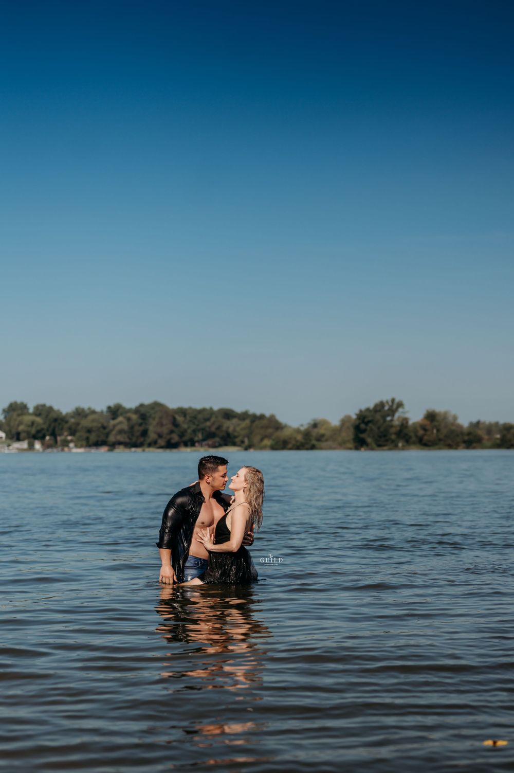 Couple standing chest-deep in a lake sharing a romantic moment under a clear blue sky.