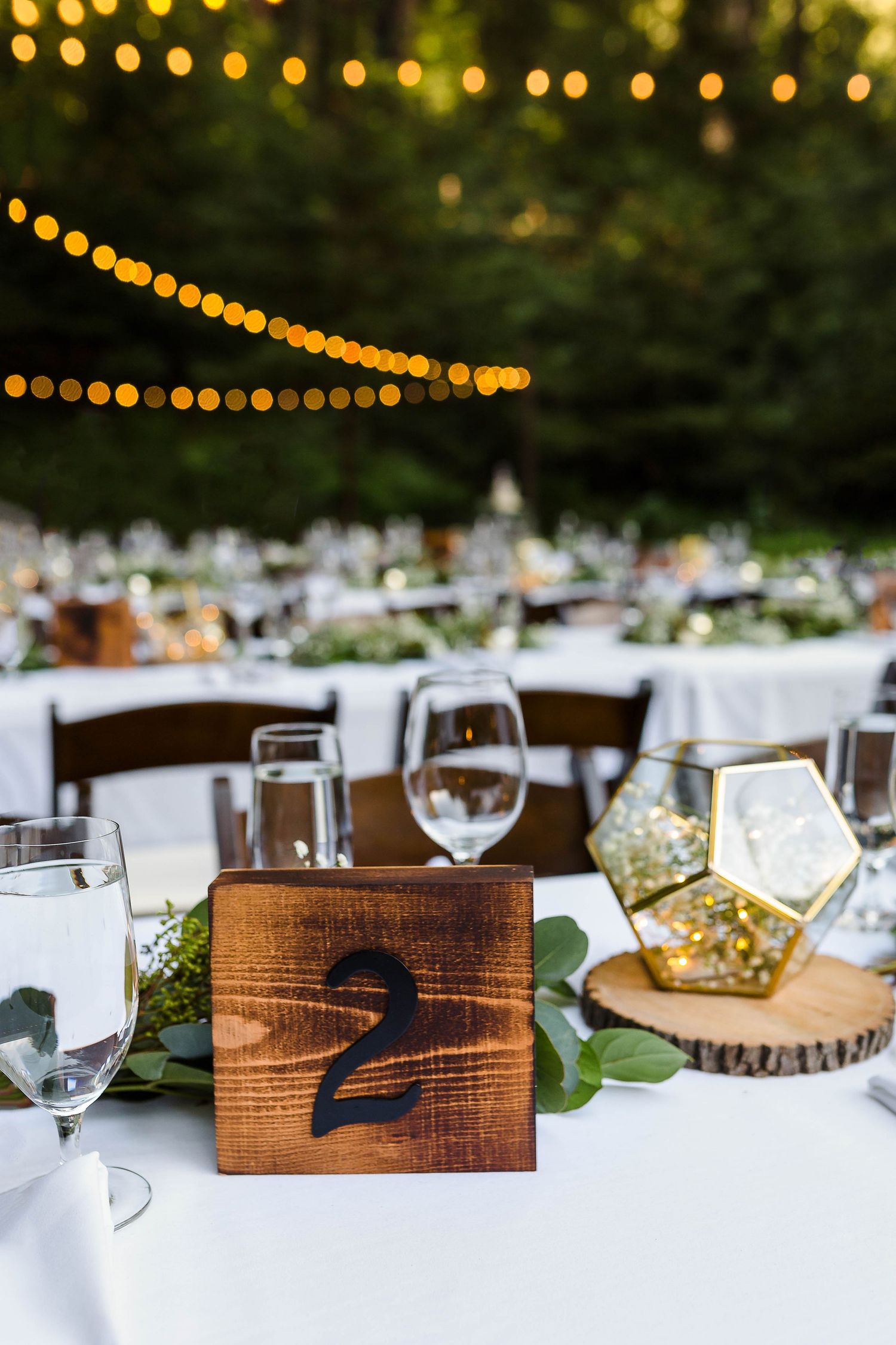 Rustic wooden table number two at an outdoor wedding reception with string lights and wine glasses.