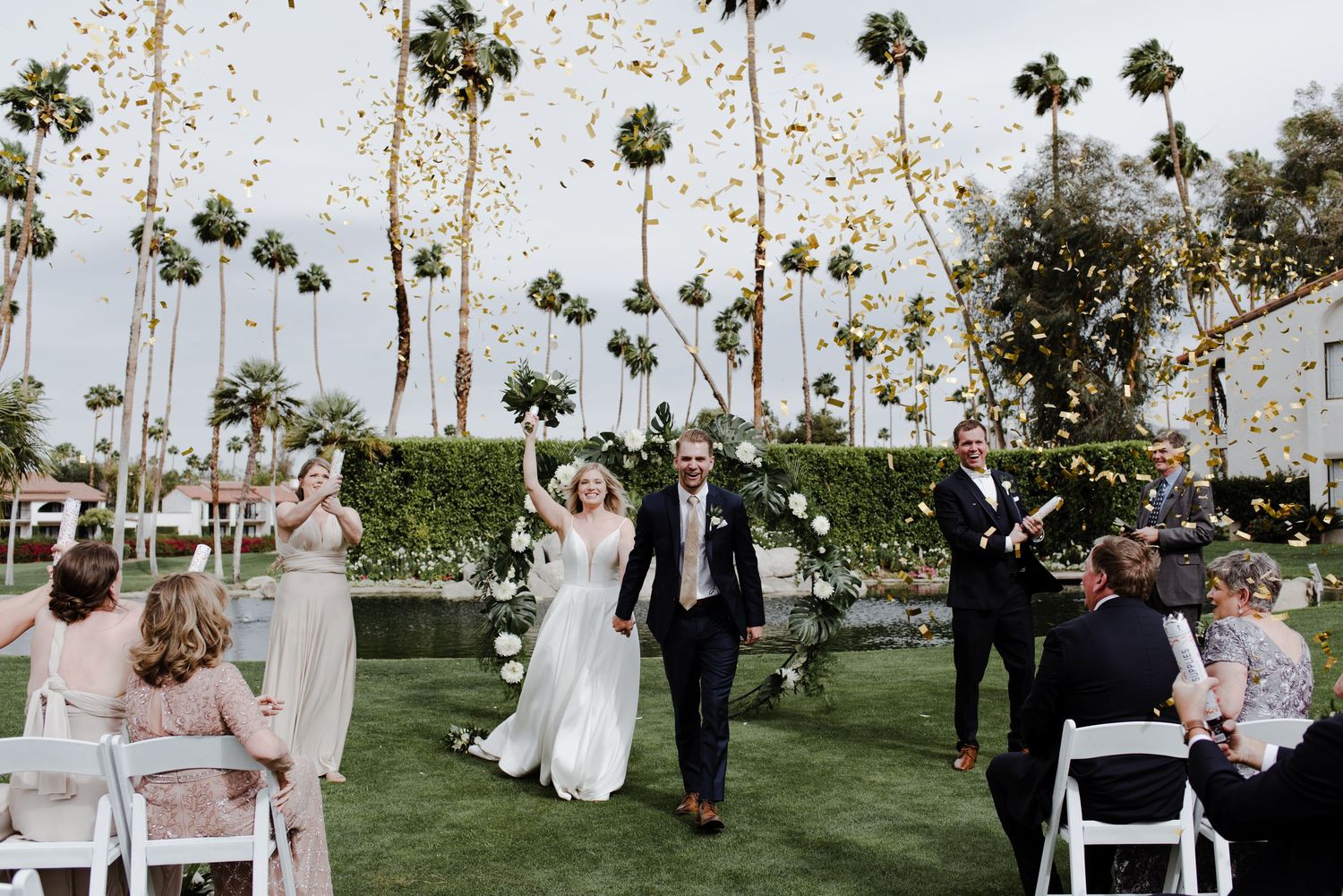 Bride with bouquet raised and smiling groom walk up the aisle from the alter, celebrating as gold confetti floats above