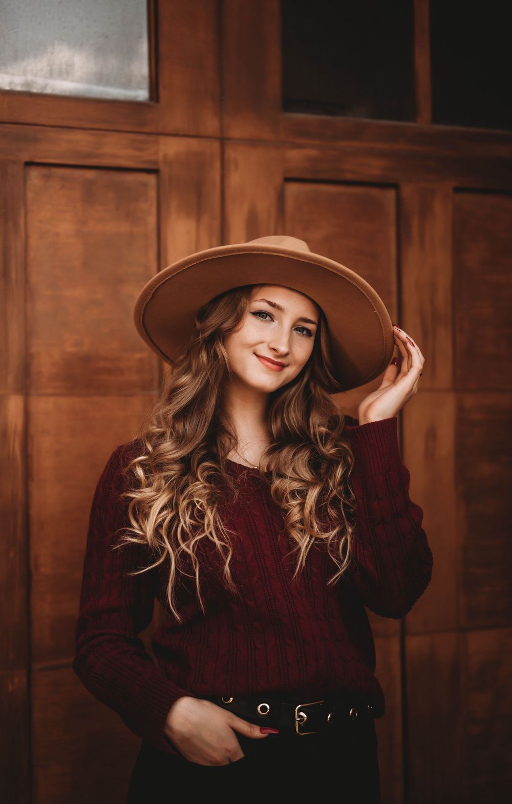 Fashionable autumn portrait featuring burgundy sweater and wide brim brown hat against wooden paneled wall.