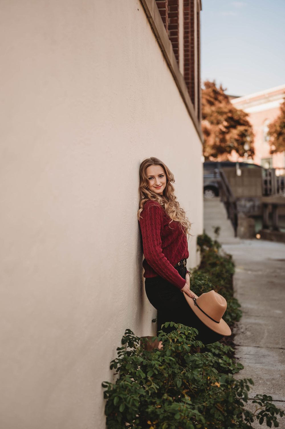 Fall fashion portrait by white wall with burgundy sweater and tan hat next to green foliage.
