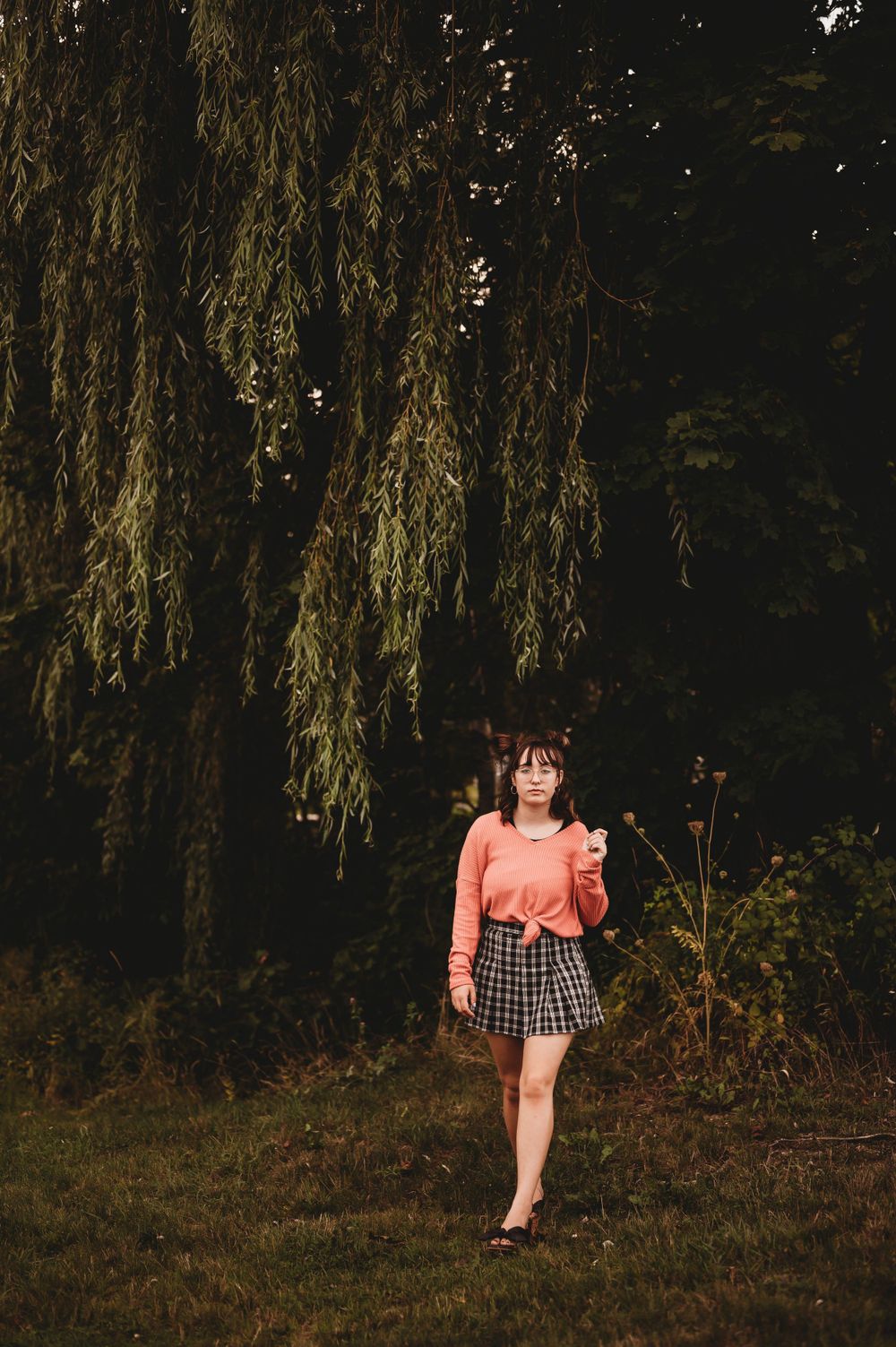 A moody portrait under a weeping willow tree wearing a pink top and plaid skirt.
