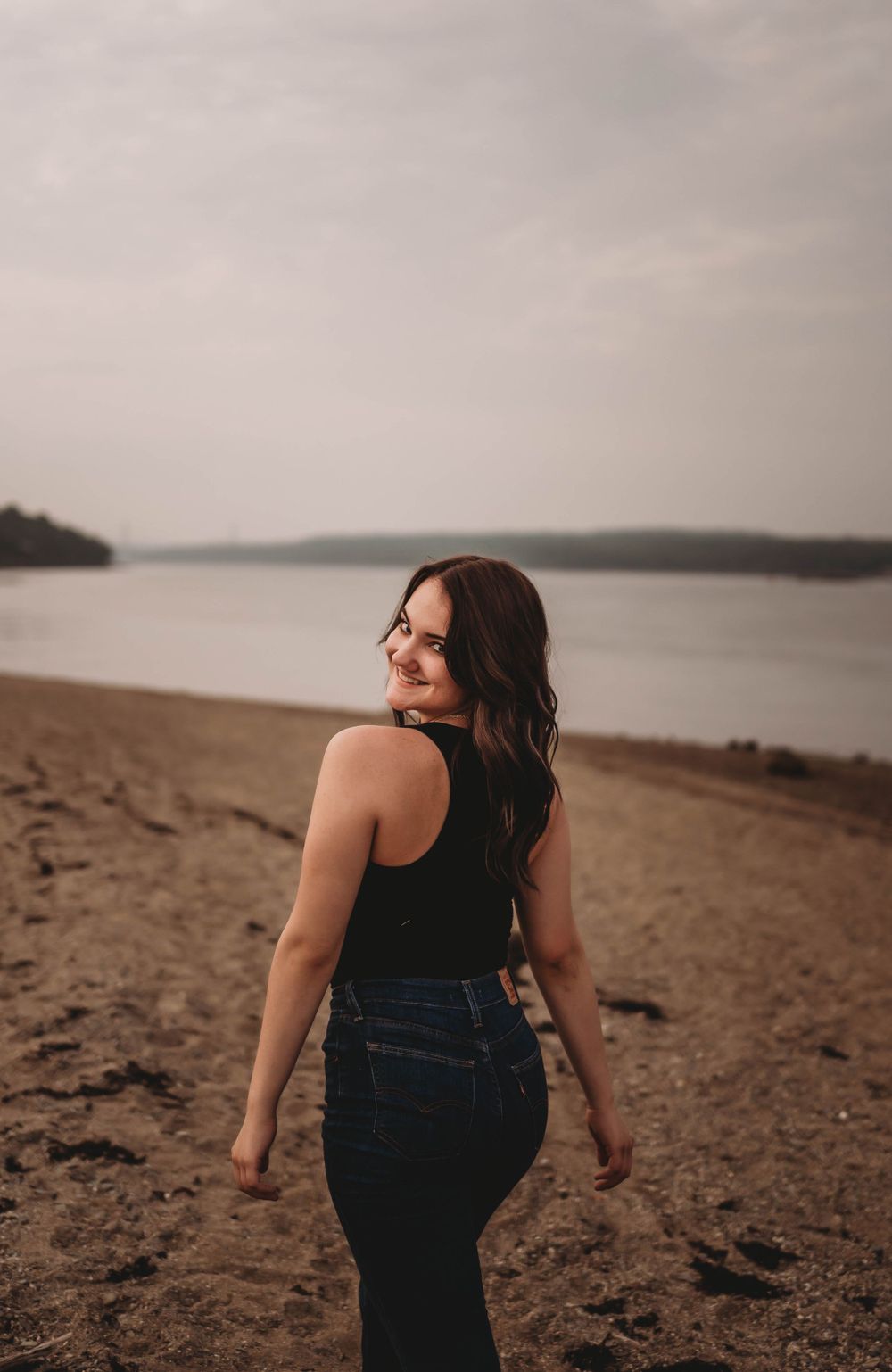 Beachside portrait in denim and black tank top with water and cloudy sky in background.