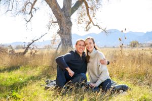 Couple sitting together under bare tree in autumn field with mountains in background.