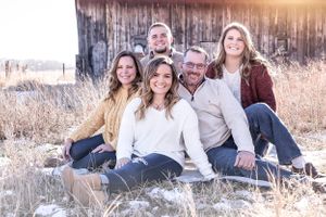 Family portrait in winter with people sitting together against a weathered barn backdrop.