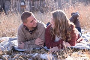 Couple lying in snowy grass having an intimate moment near a rustic barn.