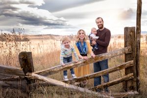 Family portrait on rustic wooden fence with dramatic cloudy sky and field background.