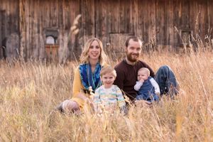 A family sits together in tall golden grass near a rustic wooden barn on a sunny day.