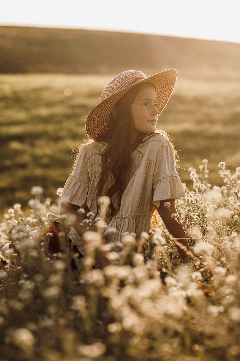 Woman in straw hat and white dress sitting among wildflowers at golden hour.