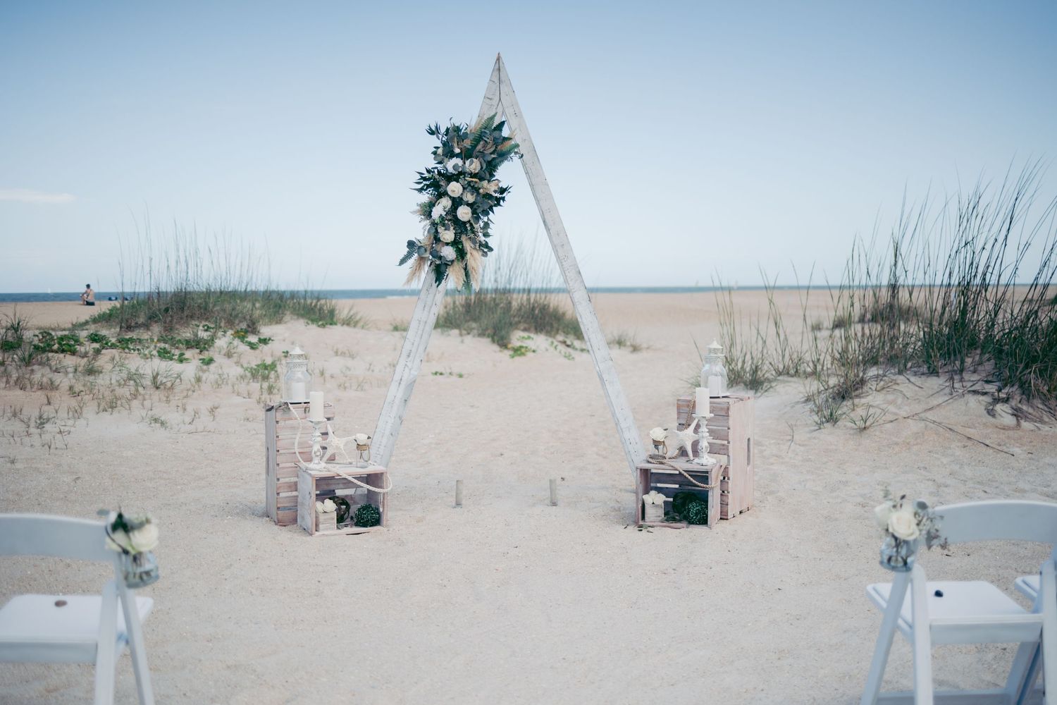 Beach wedding ceremony setup with white chairs and aisle decorations on sandy shore.