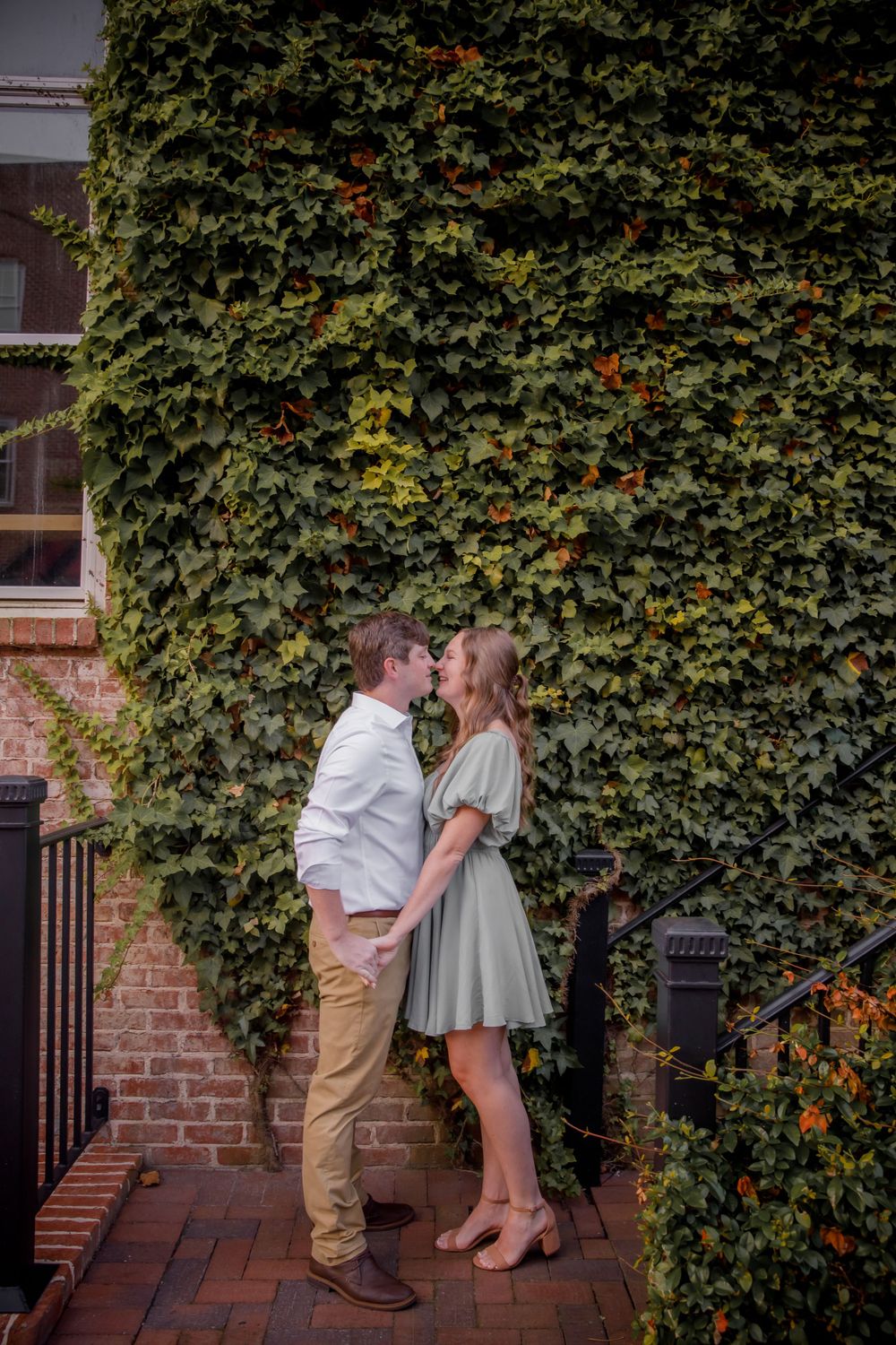 A romantic couple embraces in front of an ivy covered brick wall wearing coordinated white and sage green outfits.