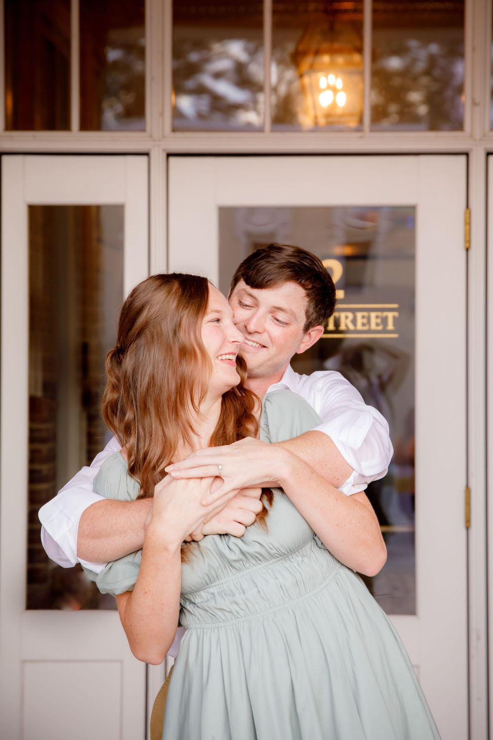 Sweet embrace captured in front of glass storefront with warm interior lighting.
