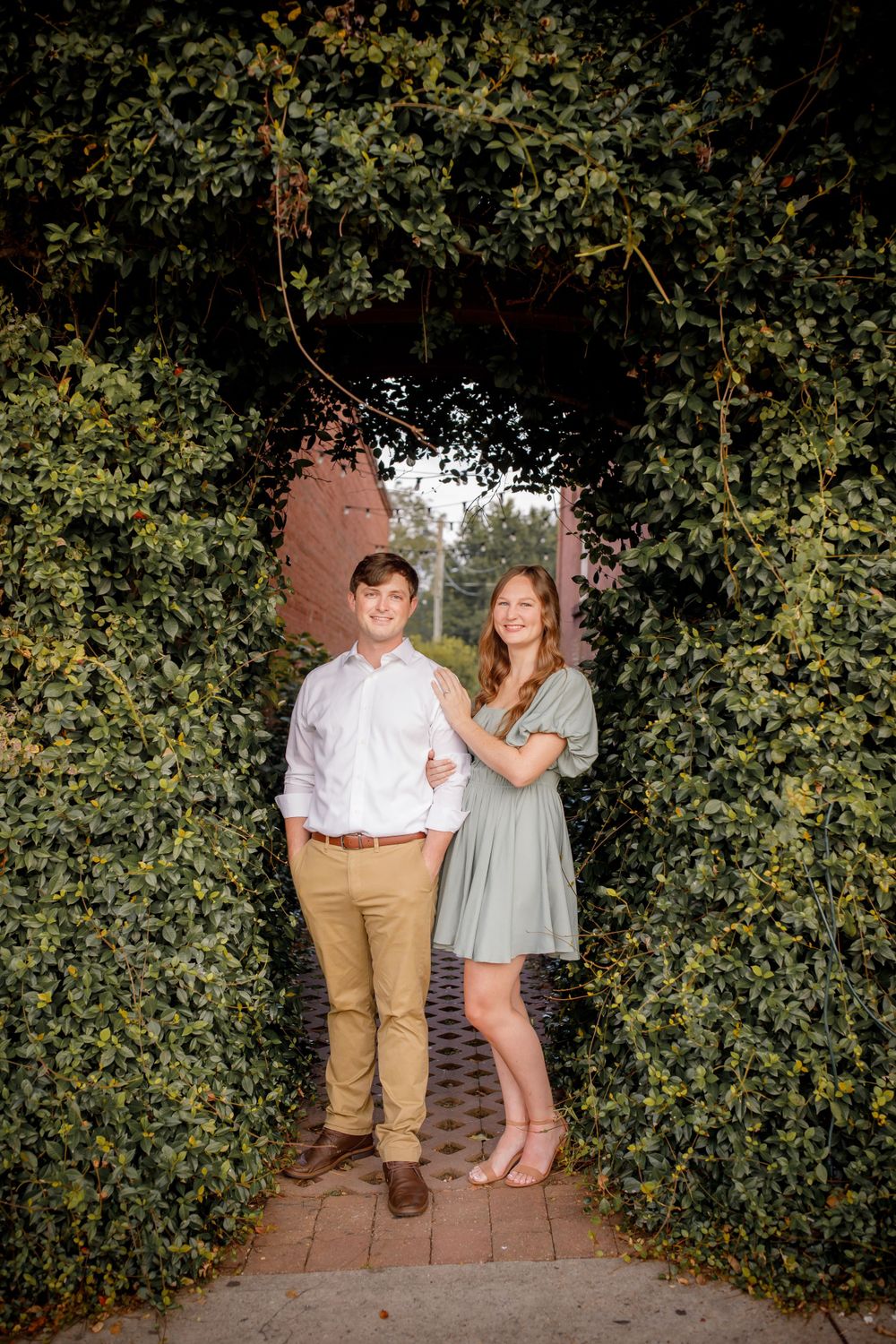 A couple poses together under a green garden archway on a brick path wearing matching casual outfits.