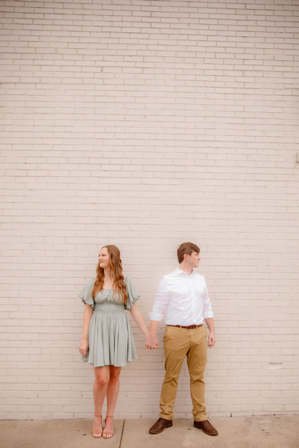 A couple holds hands while standing against a white brick wall, wearing casual attire.