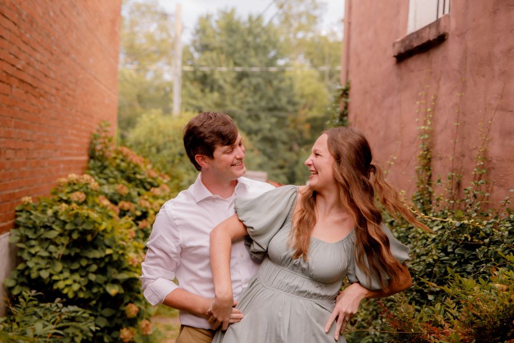 A couple shares a playful moment in an alley with brick walls and greenery.