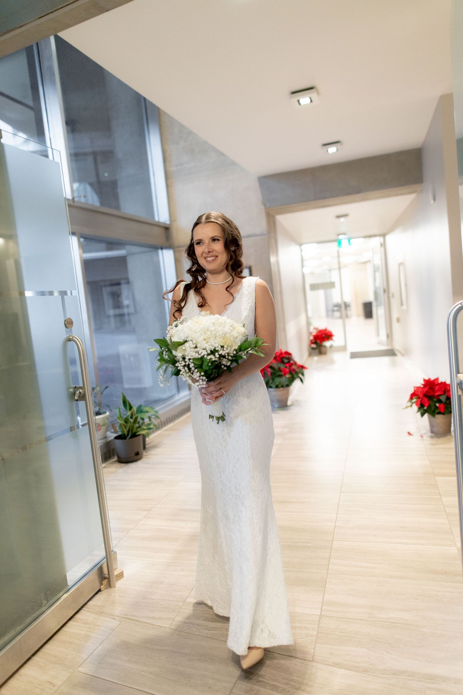 A bride in an elegant white dress walks down a modern hallway holding her bouquet.
