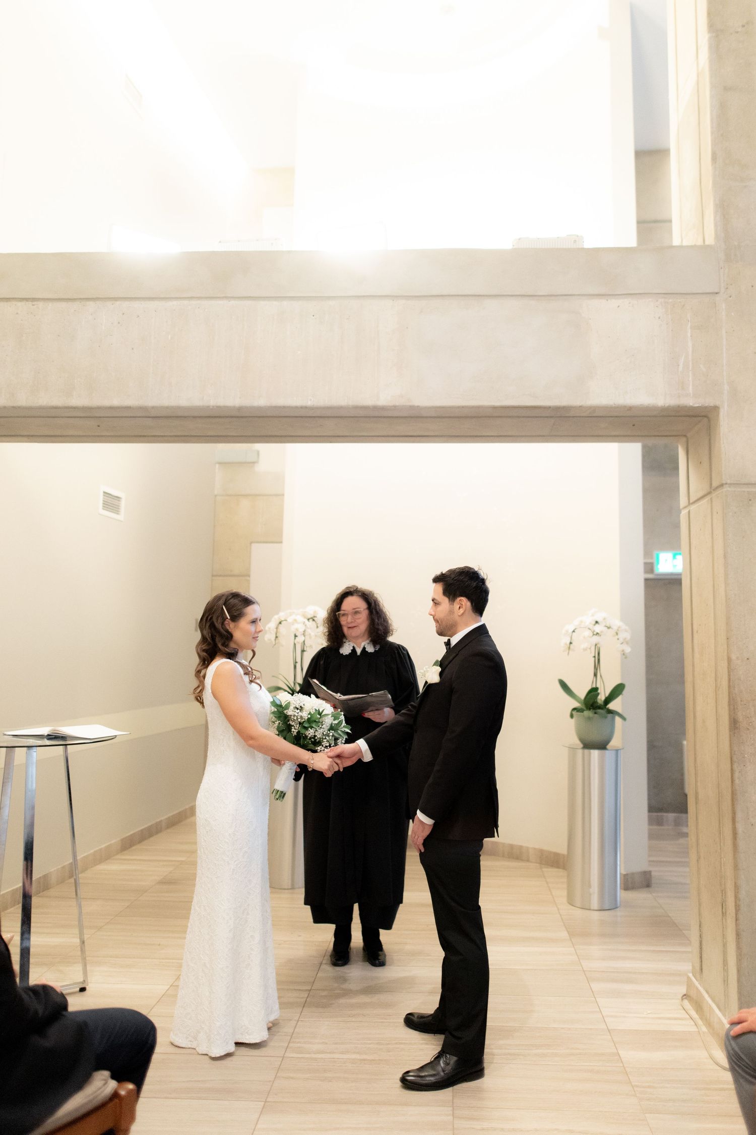 A wedding ceremony takes place in a modern minimalist venue with white walls and orchid decorations.