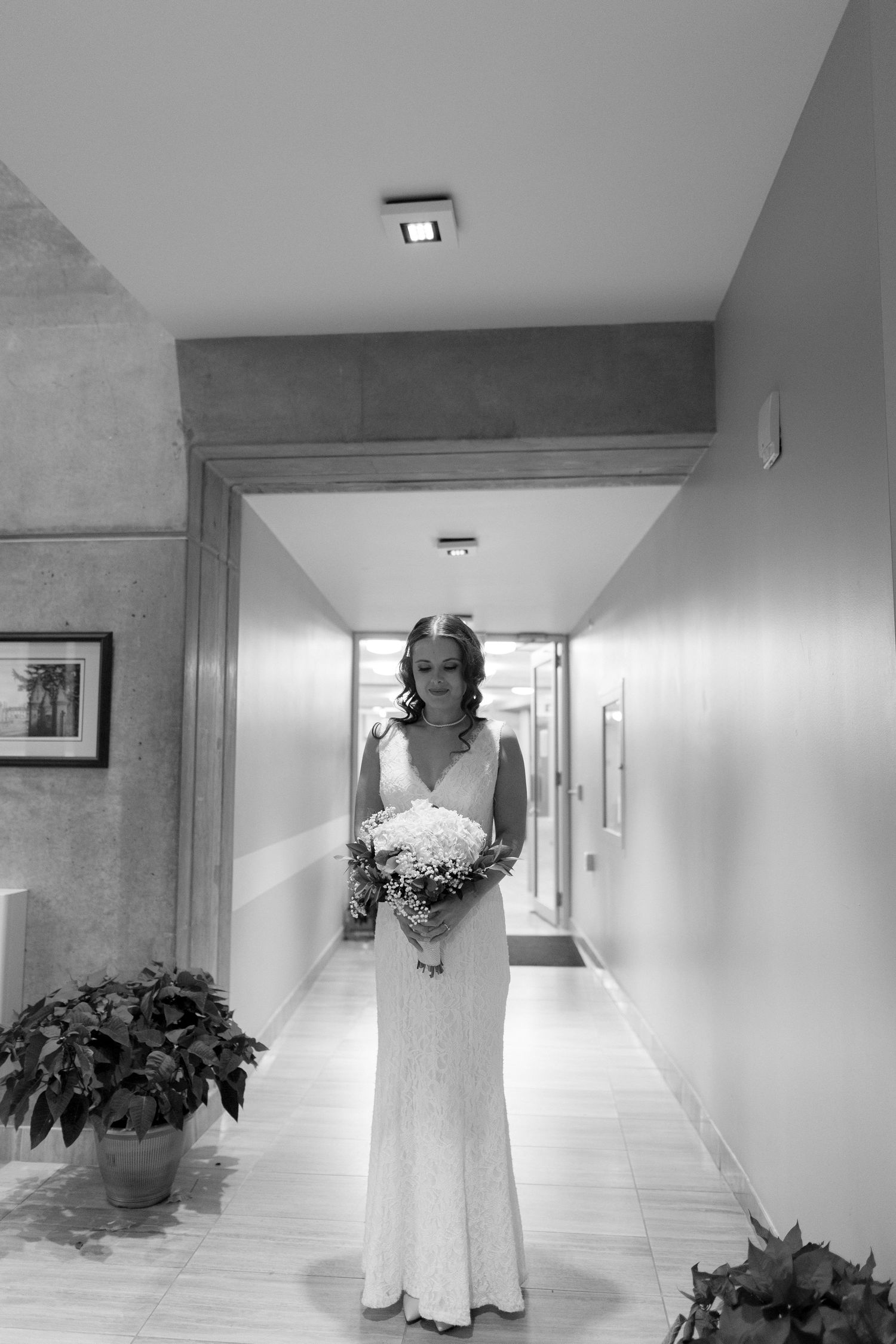 A bride in a white lace gown holds a bouquet while standing in a modern hallway.