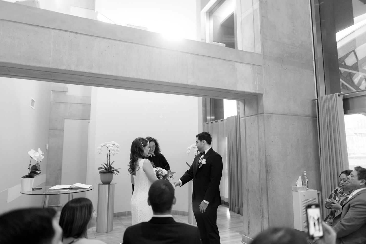 A black and white photograph captures an intimate wedding ceremony in a contemporary indoor space.