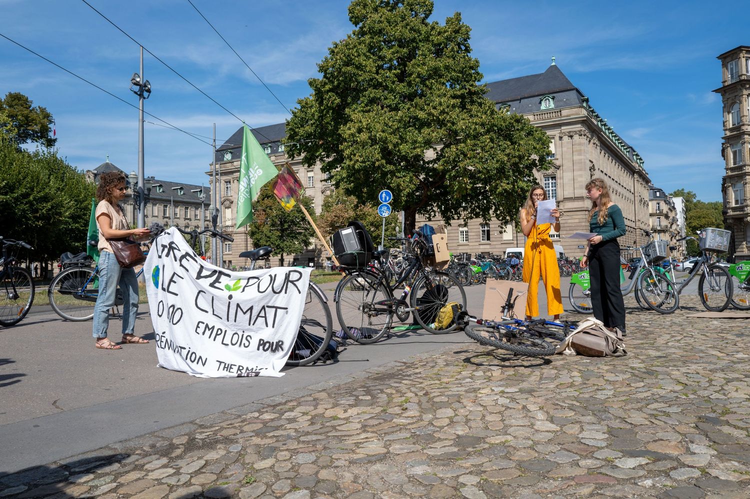 Climate activists with banners and bicycles gather on cobblestone street.