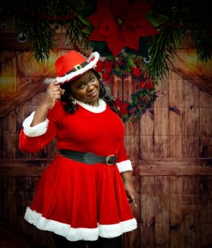 A cheerful person in a red velvet Santa dress tips their hat in front of a holiday decorated wooden wall.