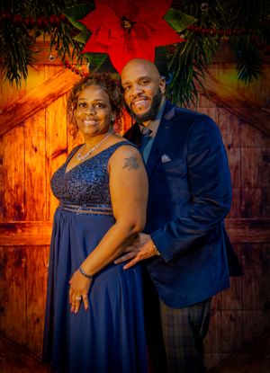 A couple in formal wear poses in front of a holiday backdrop with a large red poinsettia.
