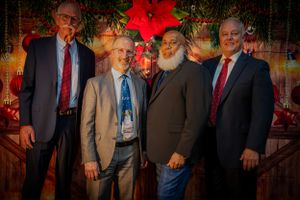 Four men in business attire pose in front of a wooden wall with holiday decorations.