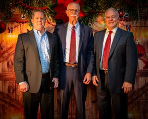Three men in business suits pose in front of a wooden wall with holiday decorations.
