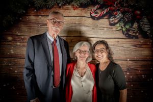 Group portrait of three people in formal wear against wooden backdrop with holiday decor.