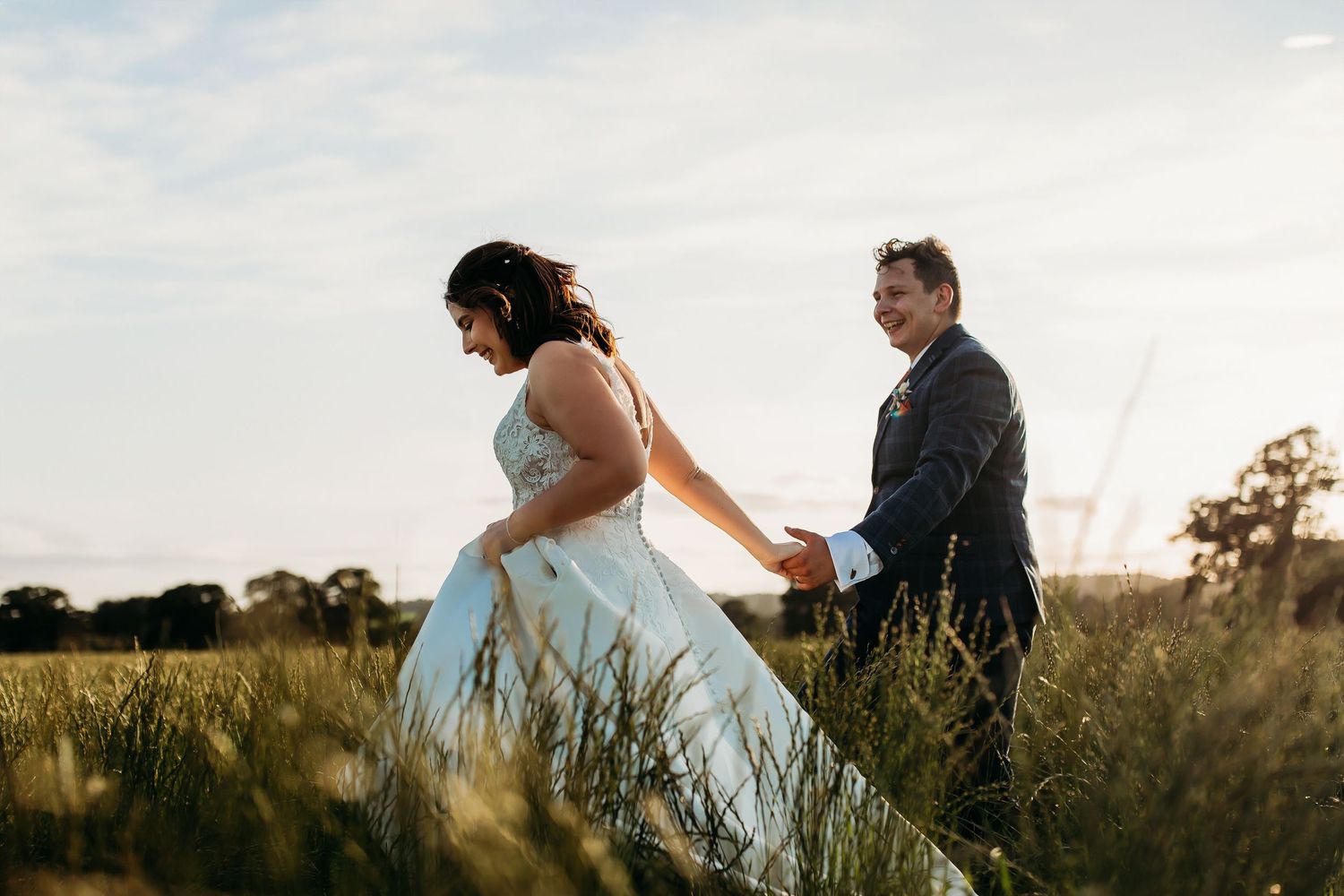 A bride in a flowing white dress walks through a golden field at sunset with a suited companion.