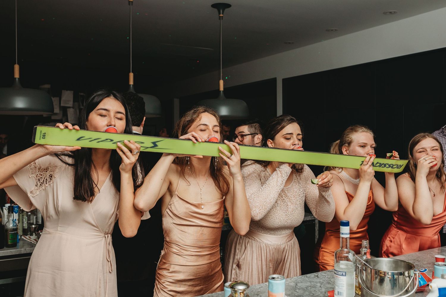 Bridesmaids in neutral-colored dresses playfully biting a green celery stick together at a reception.