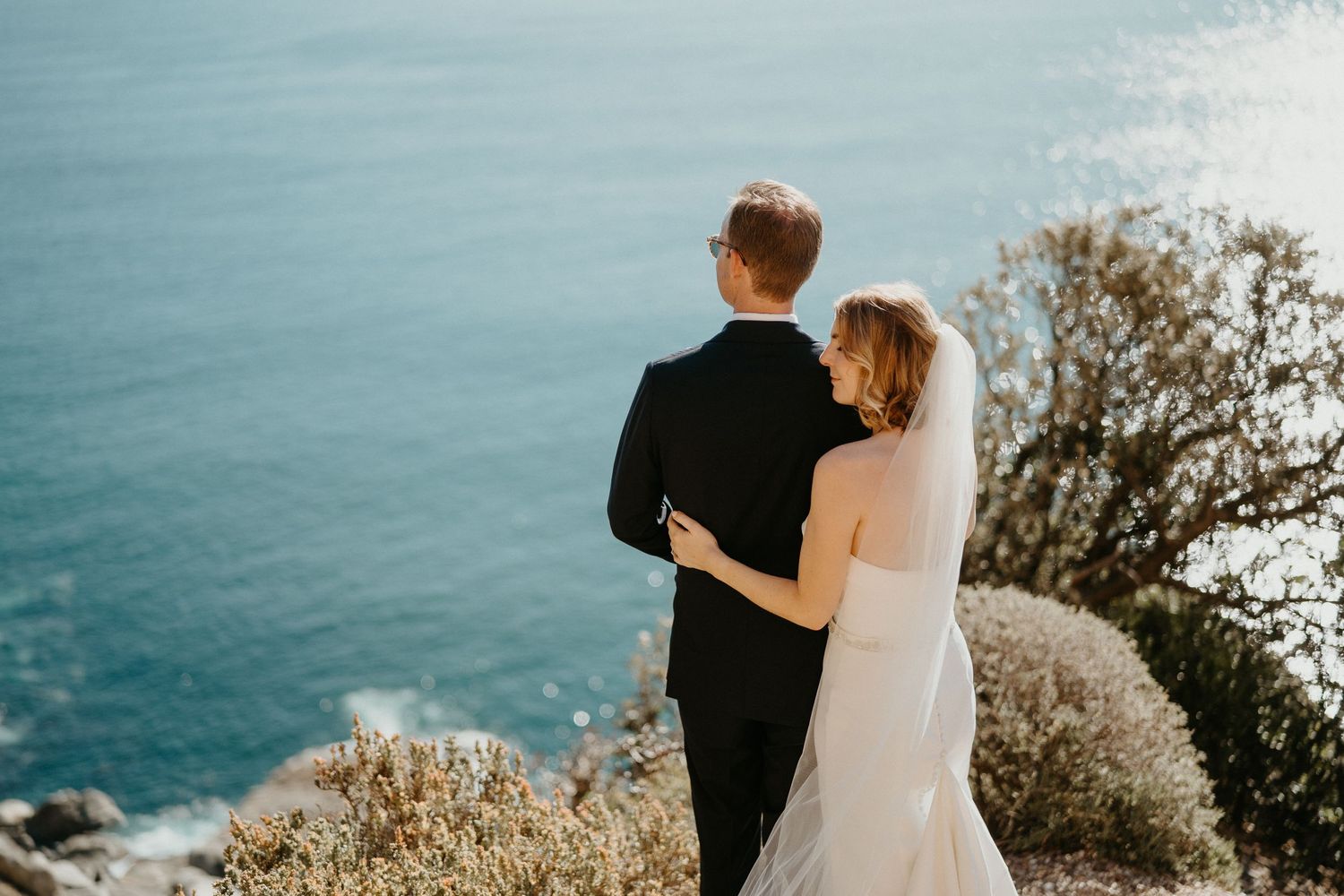 A scenic wedding photo overlooking bright blue ocean waters from a rocky cliff.