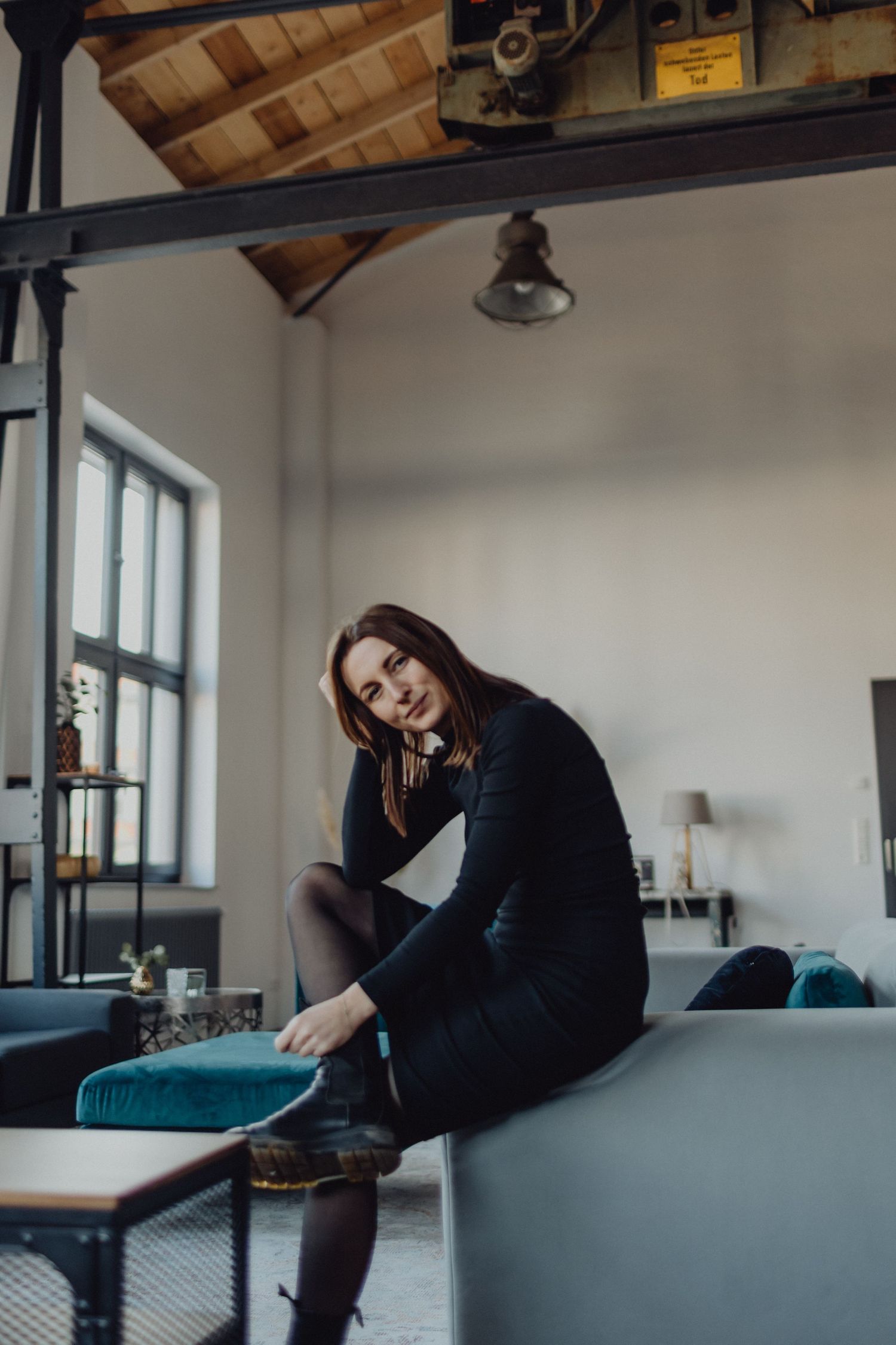 Woman in black dress sitting casually on blue couch in modern industrial loft space.