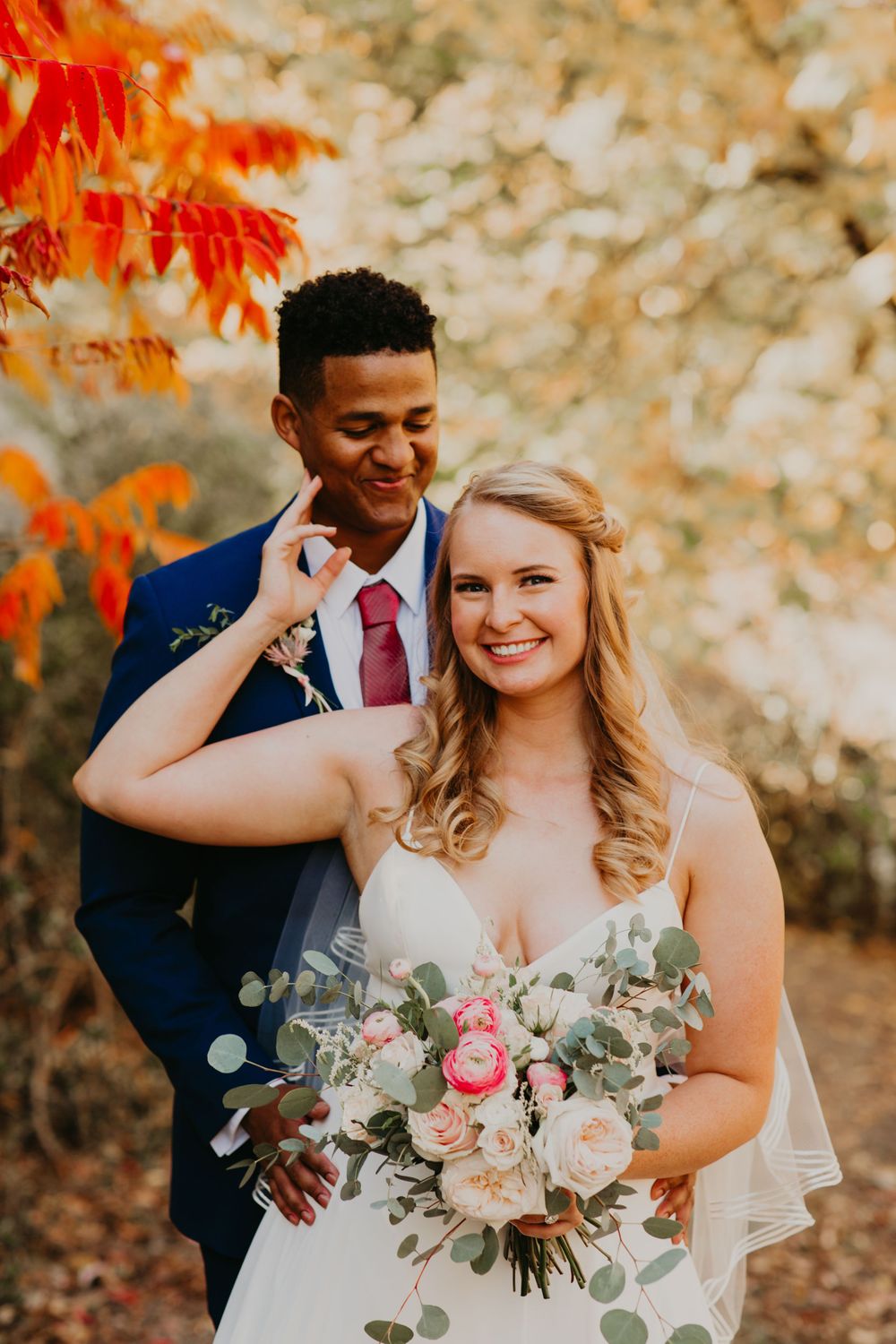 Bride and groom posing among vibrant autumn foliage, bride holding a blush bouquet.