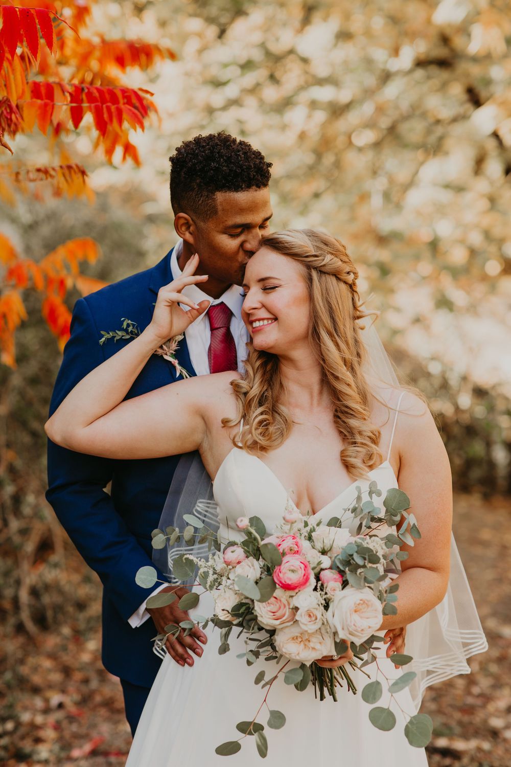 Groom kissing bride on cheek as she smiles, surrounded by fall foliage.