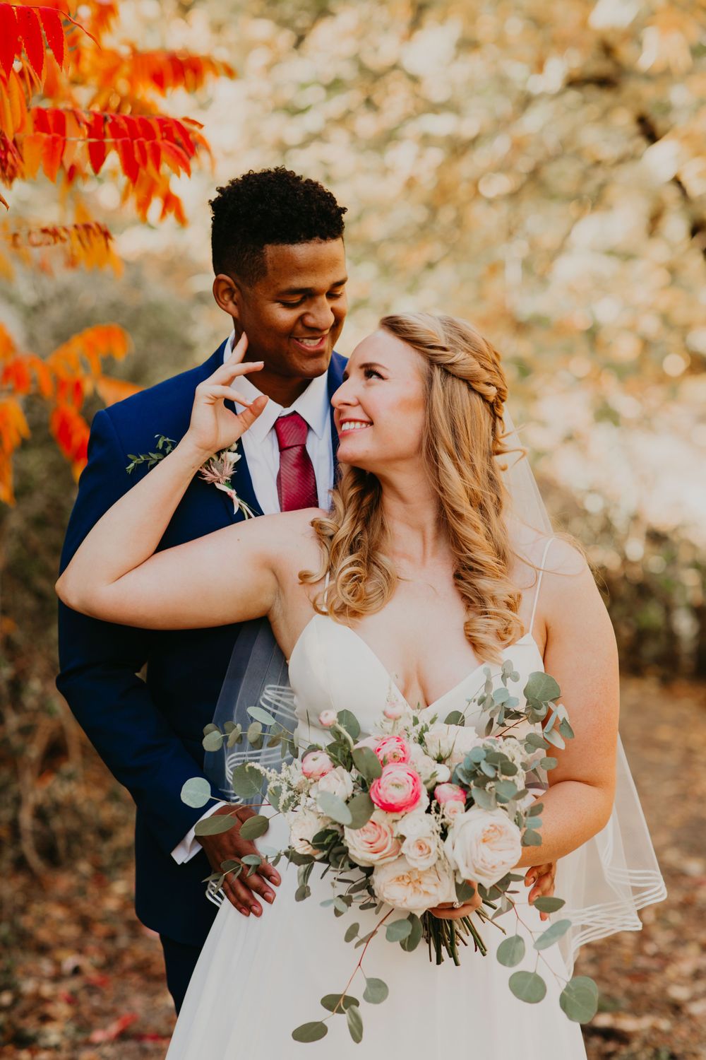 Smiling bride and groom sharing a tender moment among colorful autumn leaves.