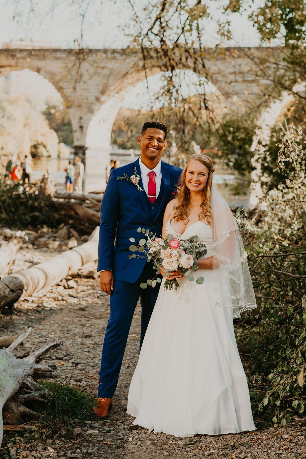 Bride and groom standing before a stone arch bridge, bride holding a blush bouquet.