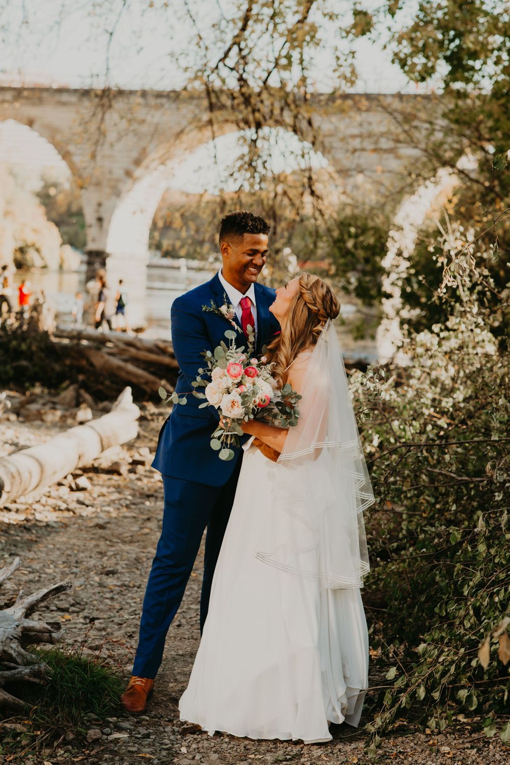 Bride and groom laughing together near a historic stone bridge on their wedding day.