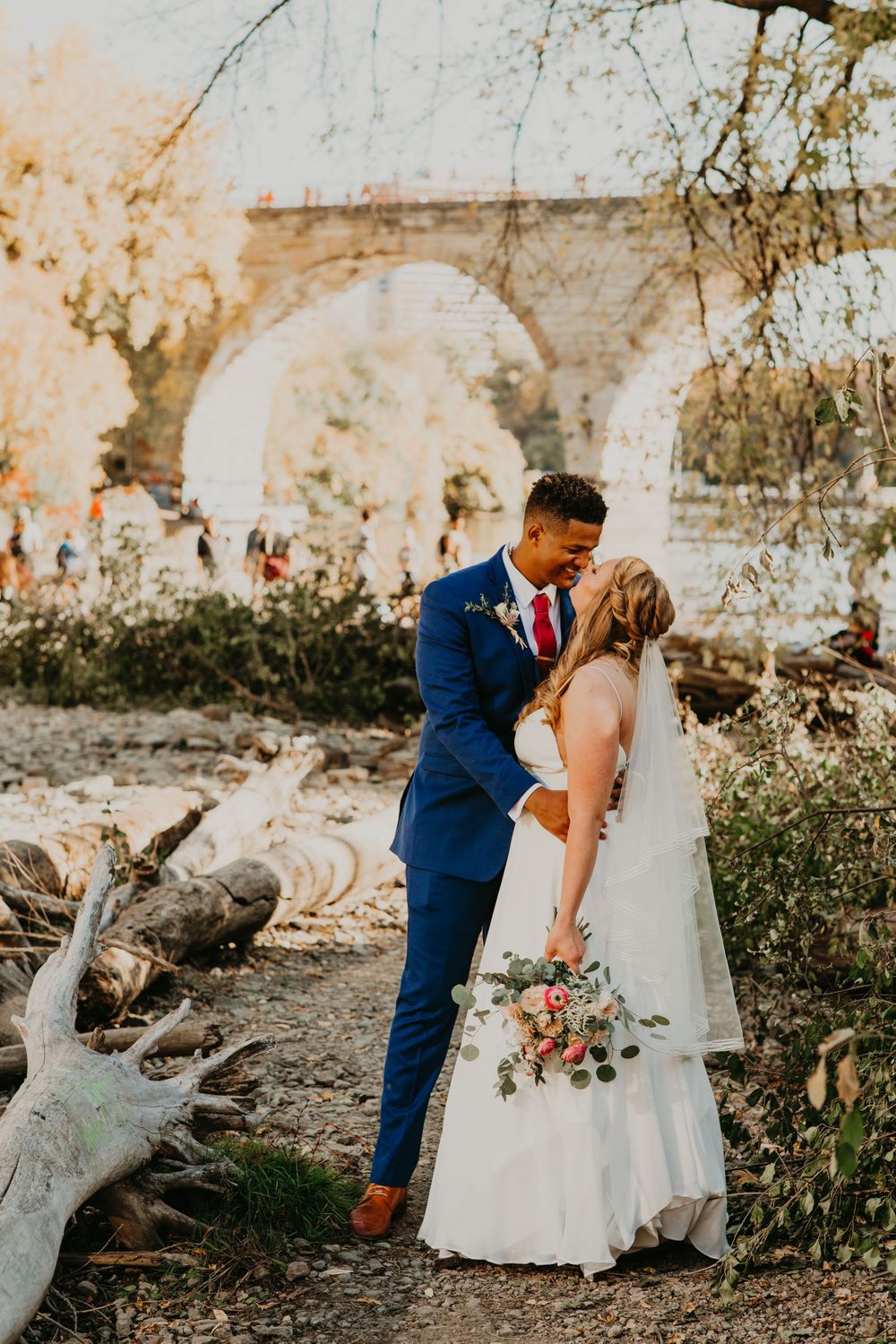 Bride and groom sharing a kiss near a scenic stone arch bridge outdoors.
