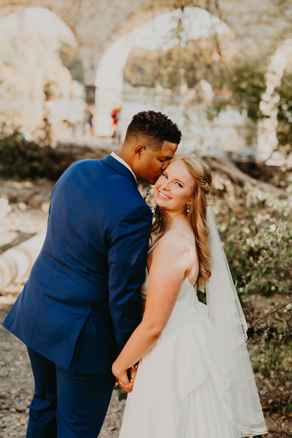 Groom kissing bride on cheek as she smiles near a historic stone arch bridge.